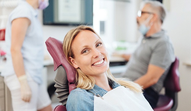A woman smiling while sitting in the dentist’s chair and the dentist and dental hygienist speak in the background